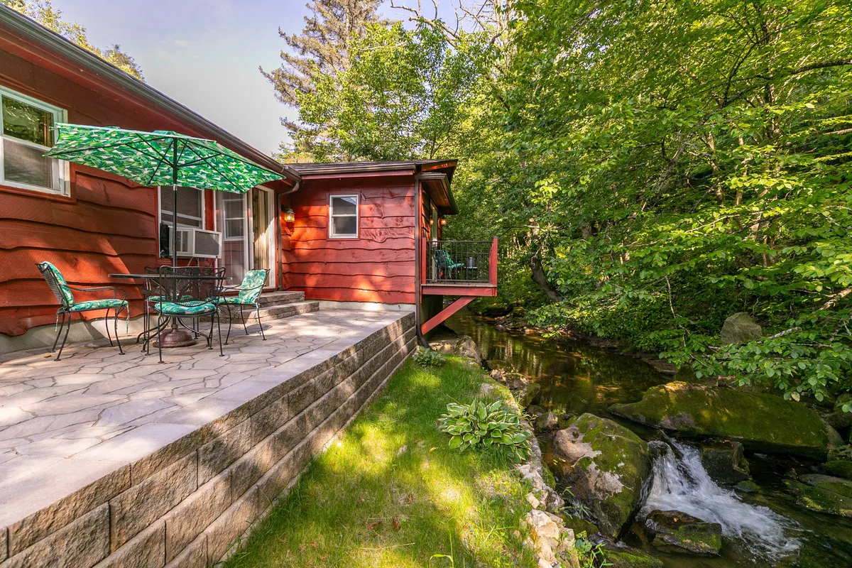This image shows a charming red wooden house with a patio set featuring a table and chairs beneath a green umbrella. The patio sits beside a flowing stream, surrounded by lush green foliage, providing a direct and tranquil connection to nature.