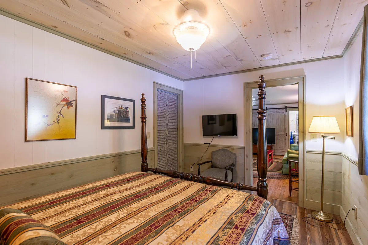 A four-poster bed dressed in soft linens and decorative pillows is the centerpiece of this inviting master bedroom. A wall-mounted flat-screen TV sits opposite the bed, flanked by a stylish lamp and tasteful artwork. Sunlight spills gently across the room, hinting at a soothing ambiance ideal for restful nights or lazy mornings.