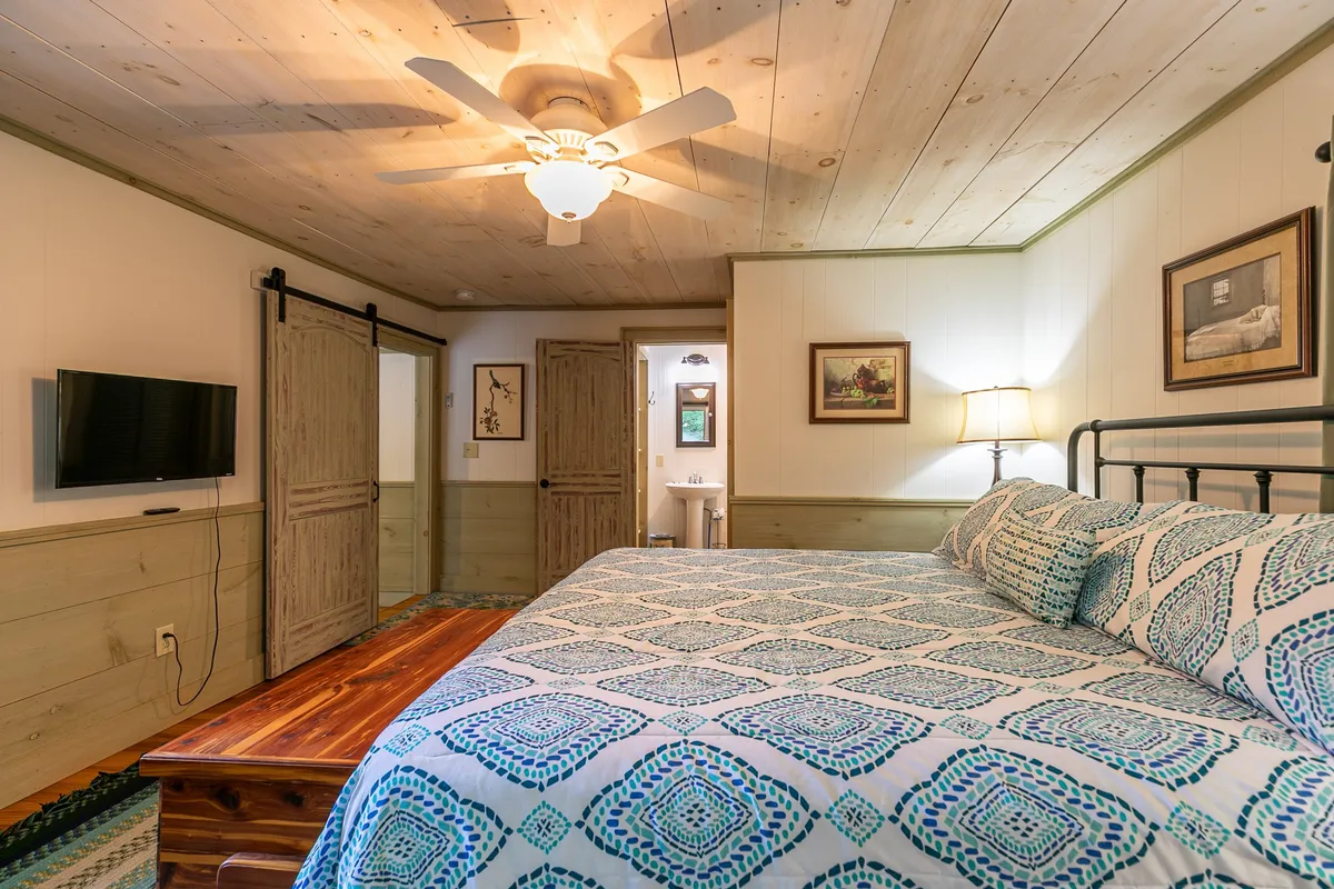 A cozy bedroom with a comfortable bed, wooden table, ceiling fan, and a wall-mounted TV. Framed artwork adds character, while natural light fills the space, creating a bright and welcoming environment ideal for unwinding after a day of adventure.