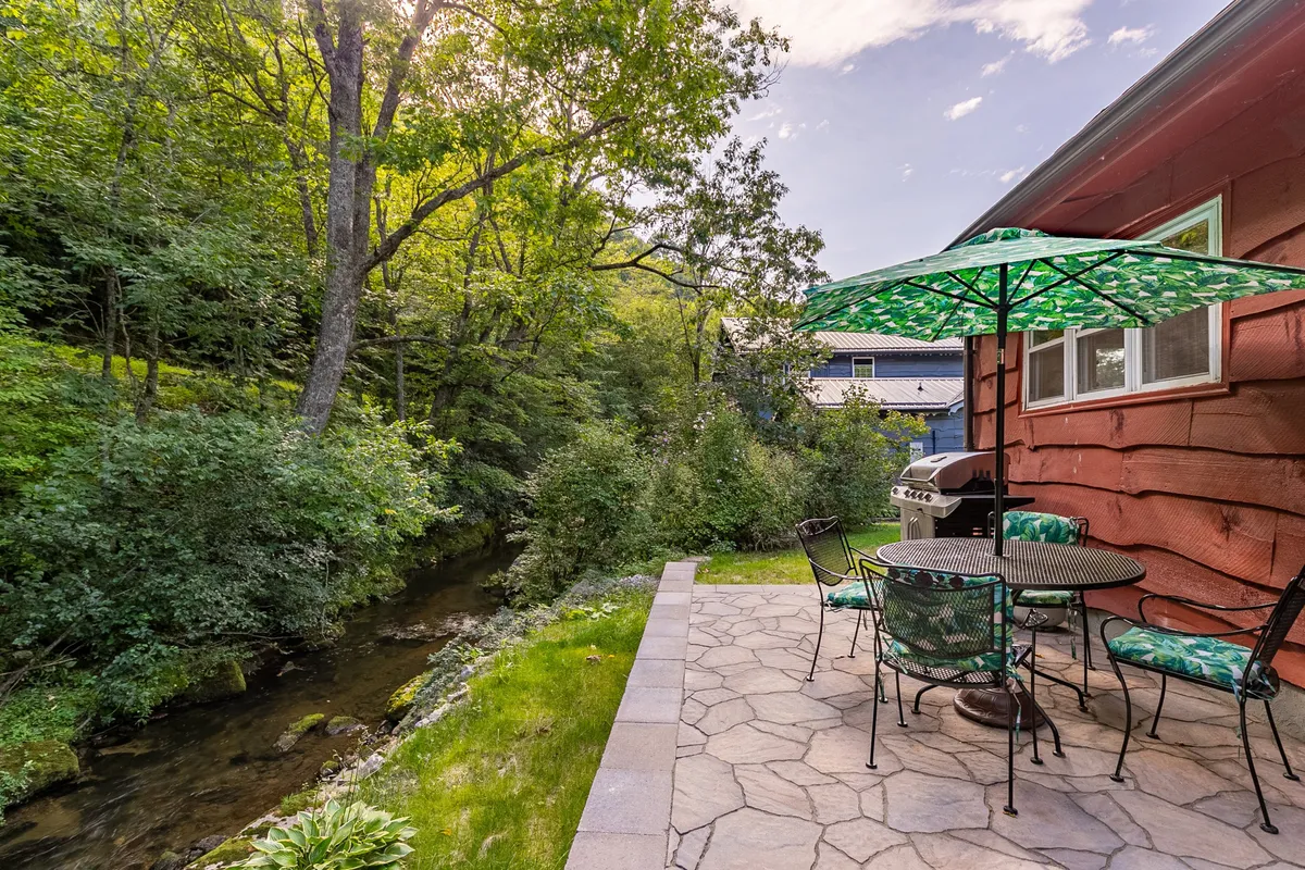 A peaceful patio scene with comfortable outdoor furniture shaded by a green umbrella, overlooking the stream and surrounded by vibrant greenery. The image evokes the idea of enjoying meals, drinks, or simply resting by the water's edge.