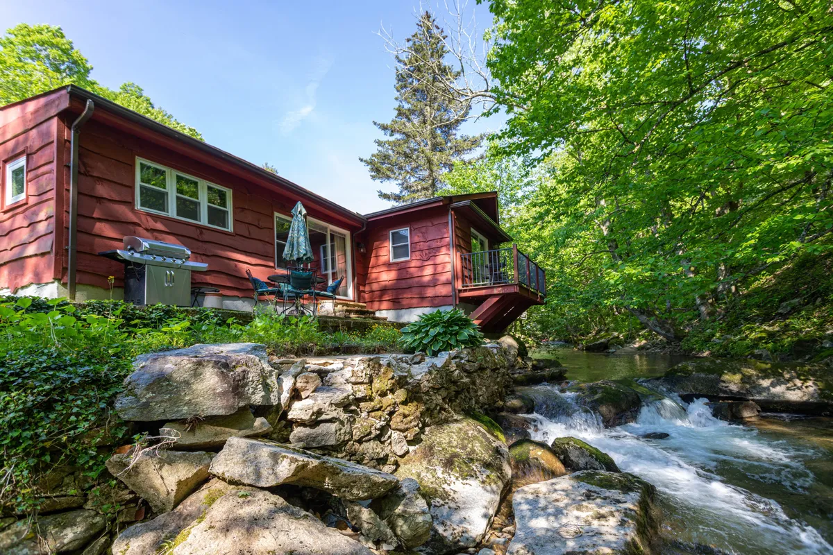 A view of the red house nestled by the stream, with a patio area featuring outdoor furniture surrounded by lush greenery. The setting shows the home’s close relationship to the water, highlighting the patio as a comfortable spot to relax and immerse in nature.