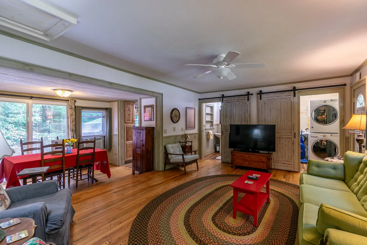 This image showcases the main living area with a green couch, red table, flat-screen TV, and wooden dining chairs. Natural light streams in through the windows, and the open-plan layout connects the seating and dining spaces, creating a sense of warmth and sociability.