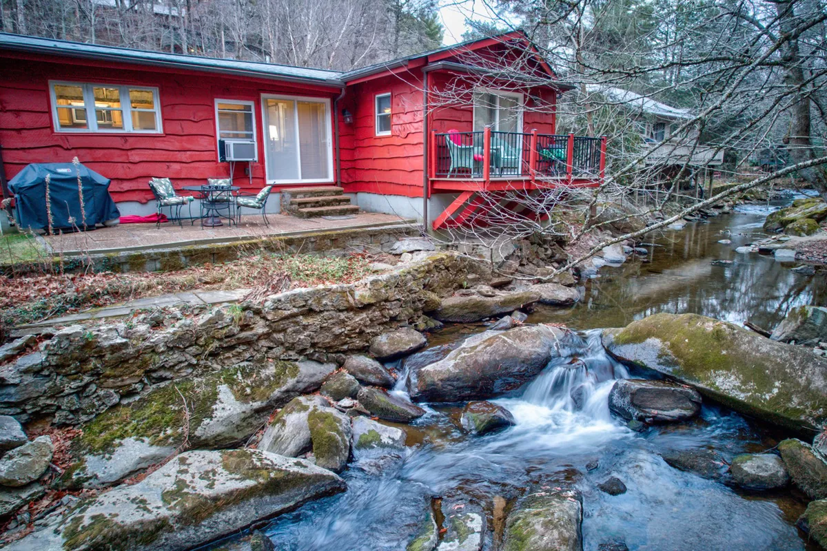 The hero image showcases a charming red single-family house nestled beside a sparkling creek, with a spacious deck overlooking the water. Surrounded by lush greenery and natural rock formations, the peaceful setting invites relaxation and connection with nature. The deck offers the perfect spot to sip coffee or watch the sunset while listening to the soothing sounds of the cascading stream.