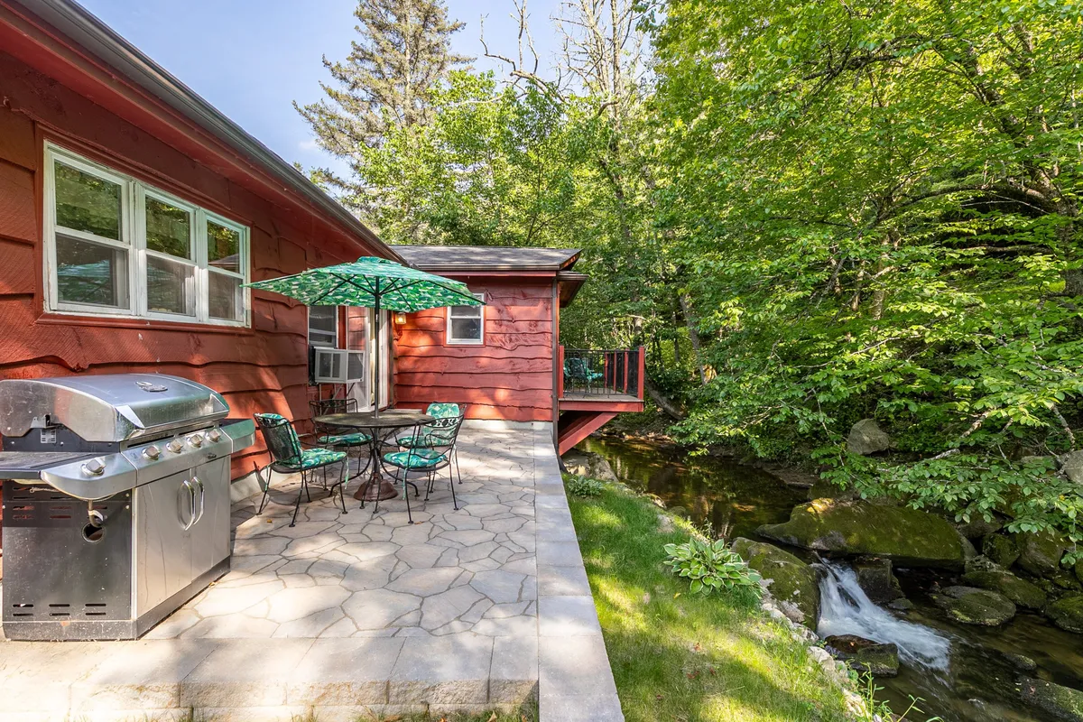 An outdoor dining setup on a deck with a grill, comfortable seating, lush greenery, and a stream nearby. The space is ideal for barbecuing, relaxing, and social gatherings, with immediate access to nature.
