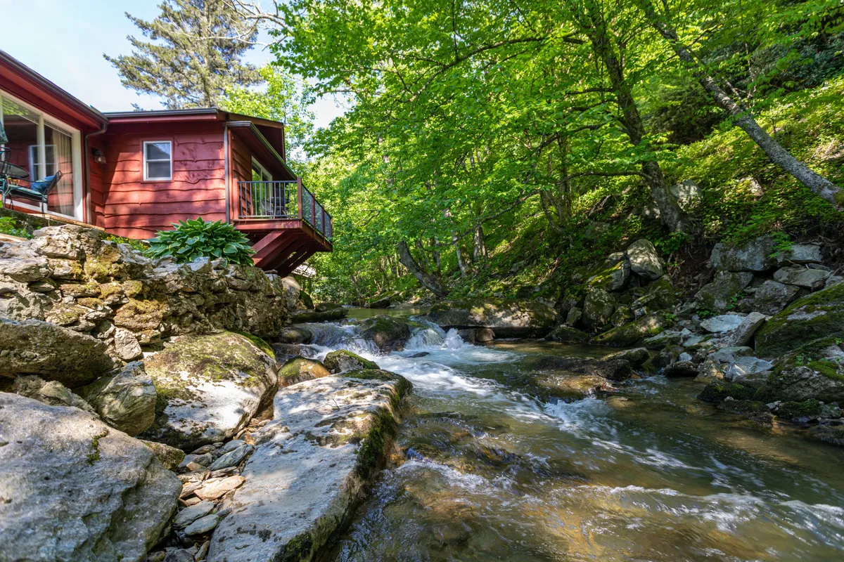 This image showcases the red house nestled beside a flowing creek, surrounded by lush greenery. The peaceful setting is accentuated by the presence of the water, the house’s inviting porch, and the natural landscape that extends beyond the property.