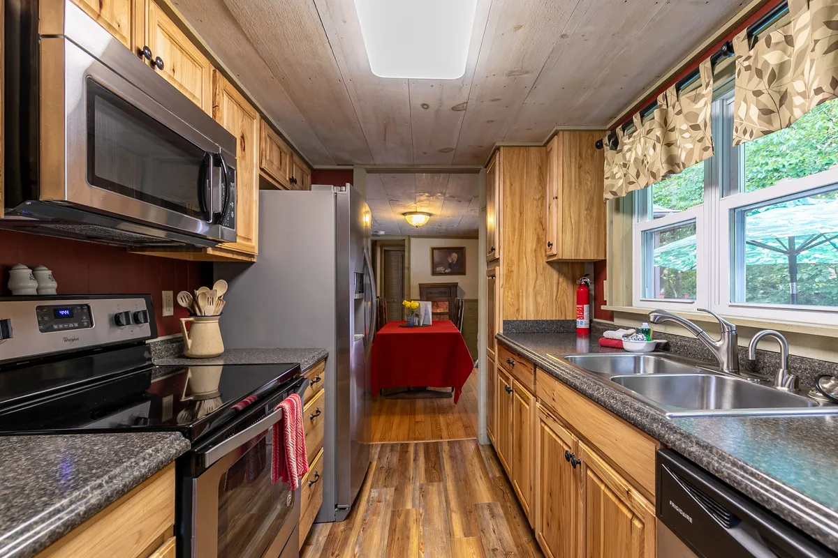 A warm, inviting kitchen features gleaming stainless steel appliances, rich wooden cabinetry, granite countertops, and a window letting in natural light. The image also offers a view into the adjoining dining area, creating a sense of flow between the spaces.