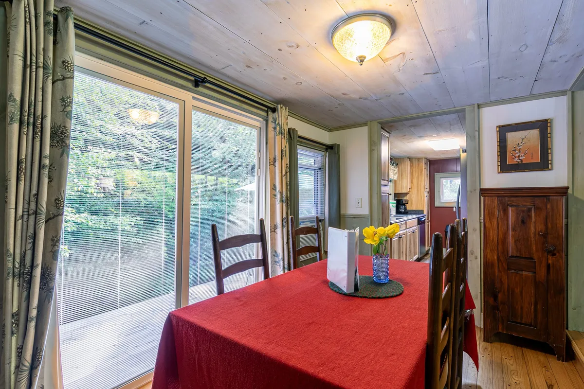A cozy dining area with a table set for six, adorned with a red tablecloth and a vase of yellow flowers. Large windows with curtains bring in natural light, and a wooden cabinet adds homey charm to the space.